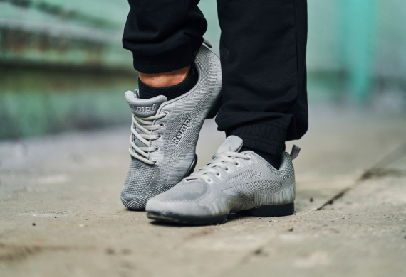 Graue Turnschuhe mit weißen Schnürsenkeln, getragen von einer Person in schwarzen Hosen, die auf einer Betonfläche steht.